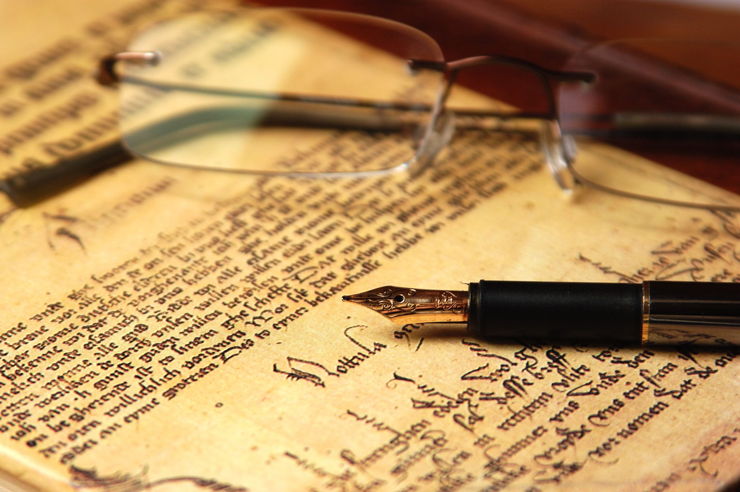 Antique parchment with cursive script, overlaid by a gold-tipped black fountain pen and a pair of glasses.