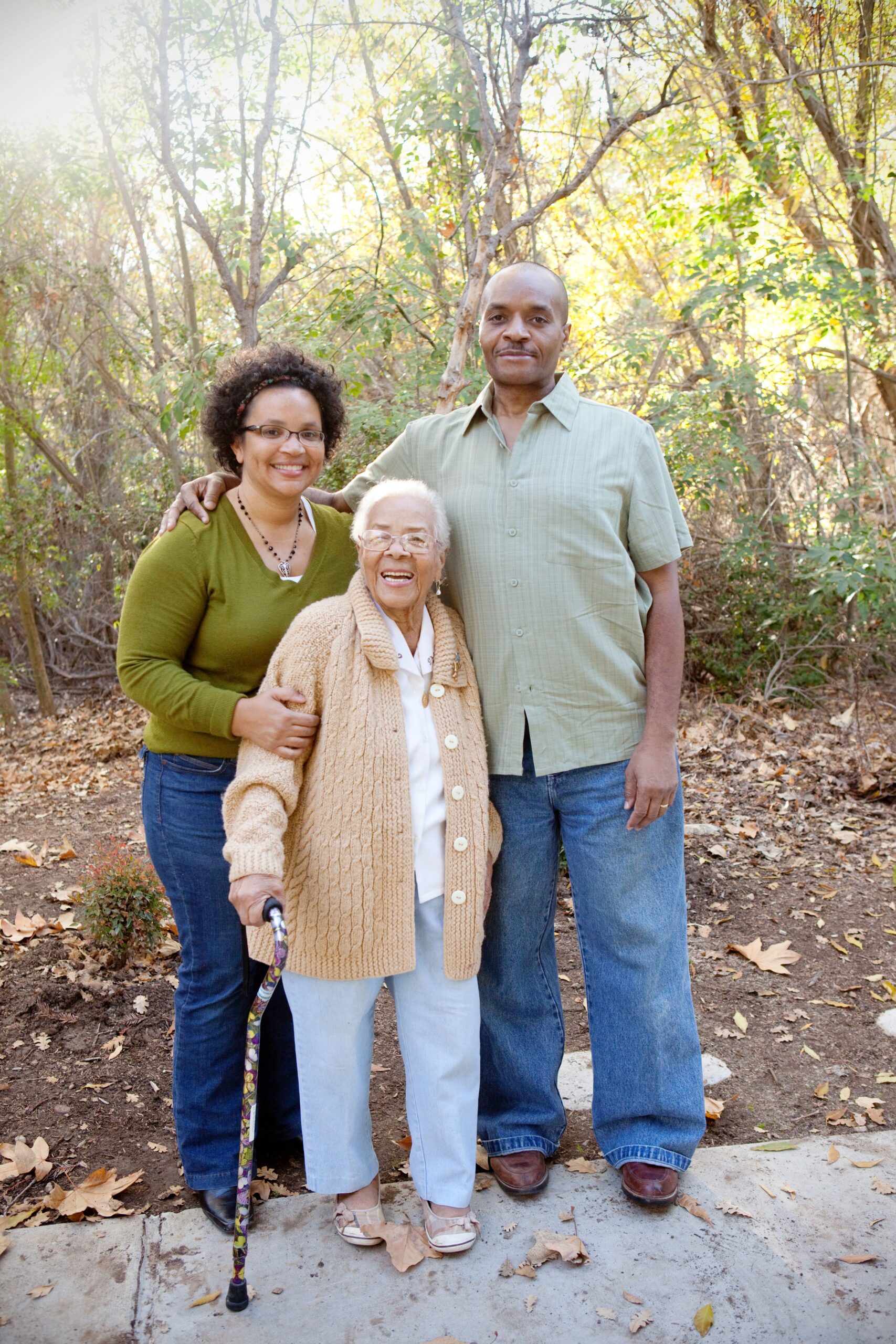 A happy elderly woman with a cane stands between two adults outdoors in a forest