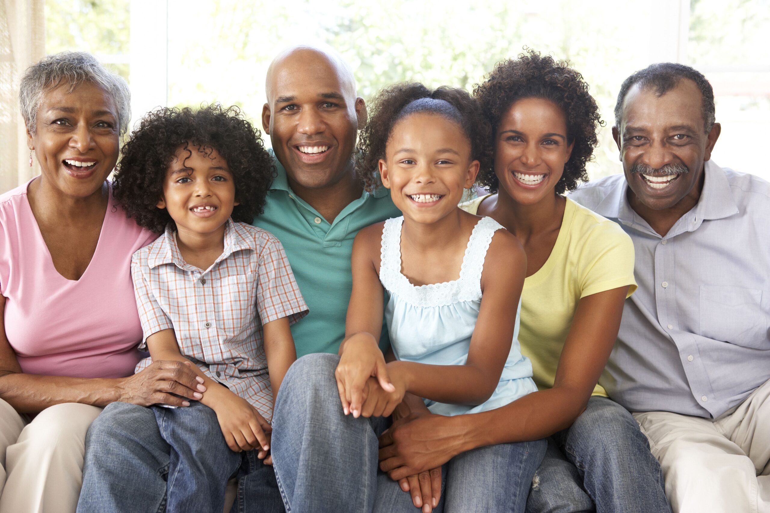 A joyful family of six sits closely on a sofa, smiling at the camera.