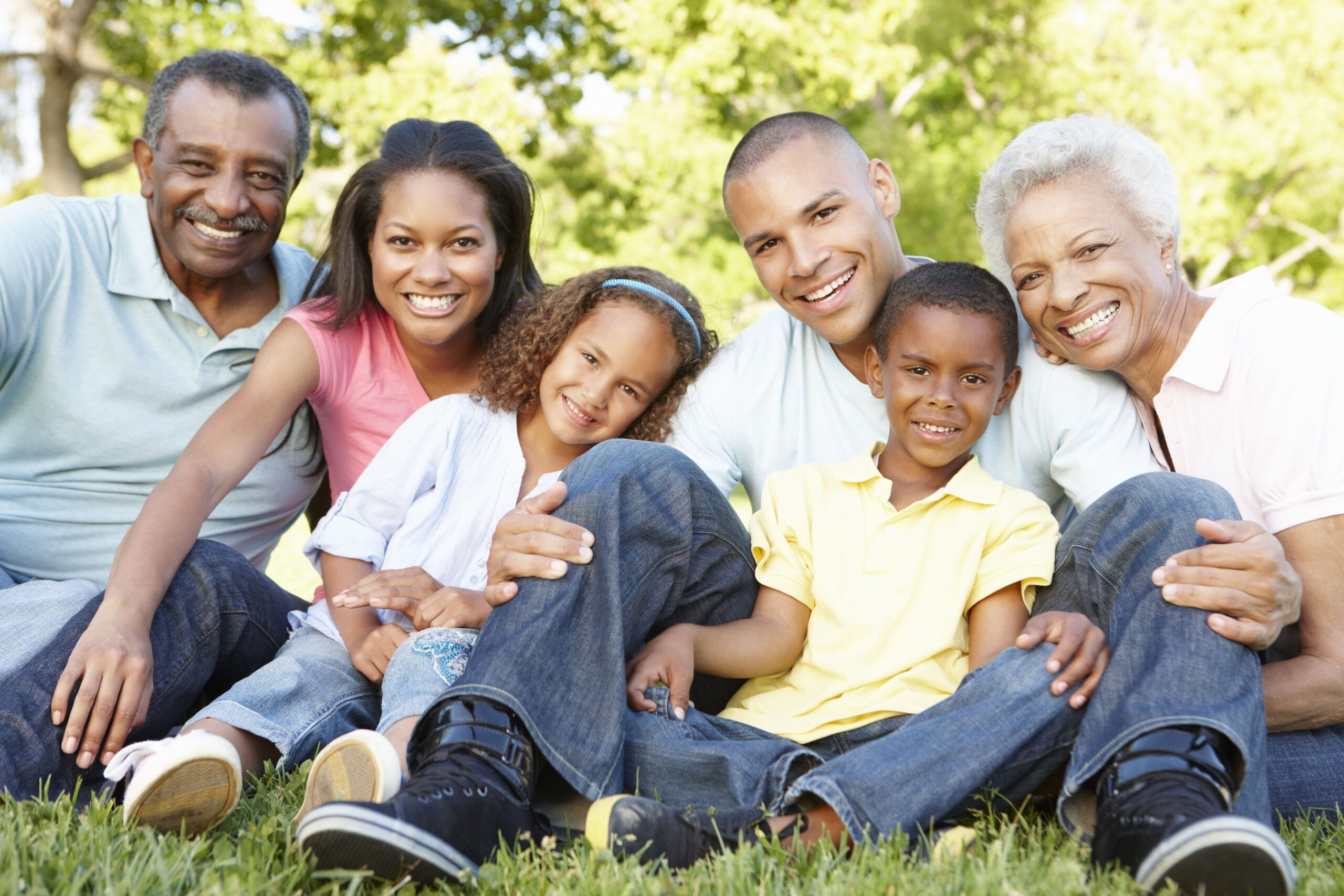 A smiling, multi-generational family sitting on grass in a park.