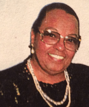 Elderly woman smiling warmly, wearing glasses, pearl necklace, and dark attire.