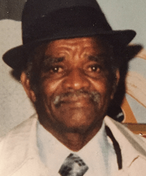 An elderly man with a gentle smile, wearing a black hat and a light-colored jacket over a white shirt and patterned tie, exudes warmth and wisdom.