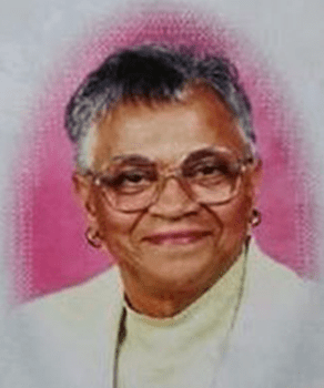 A smiling elderly woman with short gray hair and glasses wears a light yellow top and white jacket.