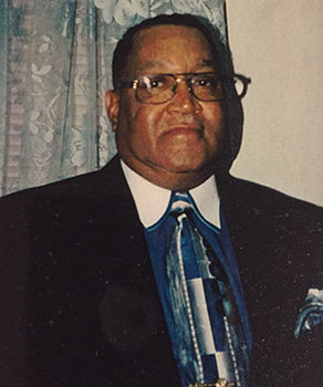 A man wearing glasses and a suit with a colorful tie stands in front of a floral curtain