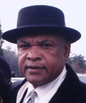 A man wearing a black bowler hat and dark coat over a suit and tie looks directly at the camera.